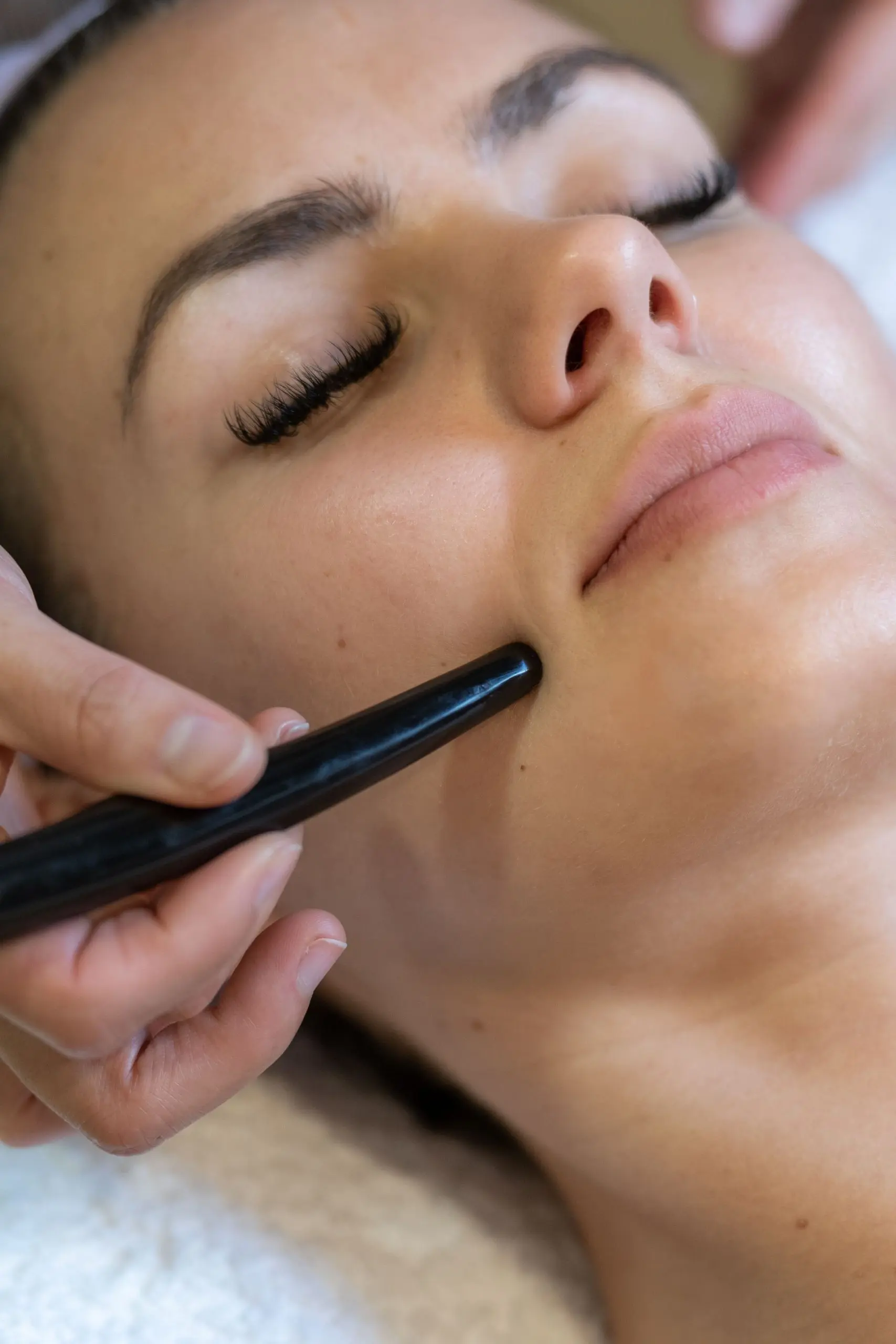A close-up shot of a person's face receiving a facial treatment. A close-up shot of a person's face receiving a facial treatment. Their eyes are closed, and a hand is using a dark tool on their cheek. The person's skin is visible, as are their eyelashes and lips. The hand holding the tool is also visible, and the background is a soft, textured surface.